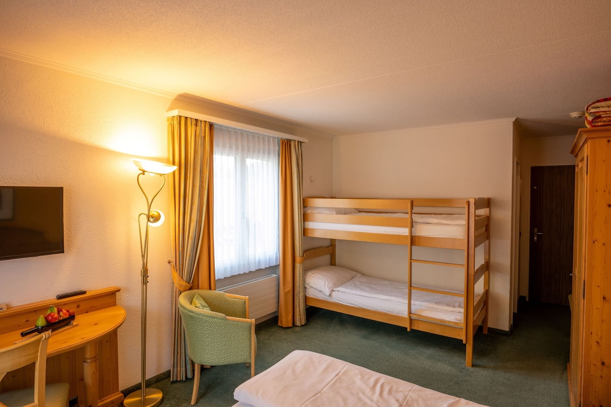 Habitación de hotel con literas, un escritorio con silla, una lámpara y una ventana con cortinas.