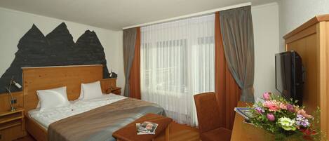 Habitación de hotel con cama, escritorio, silla, televisor y ventana con cortinas.