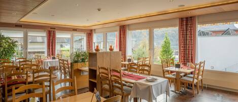 A restaurant with wooden tables and chairs, large windows with curtains, and a view of the outside.
