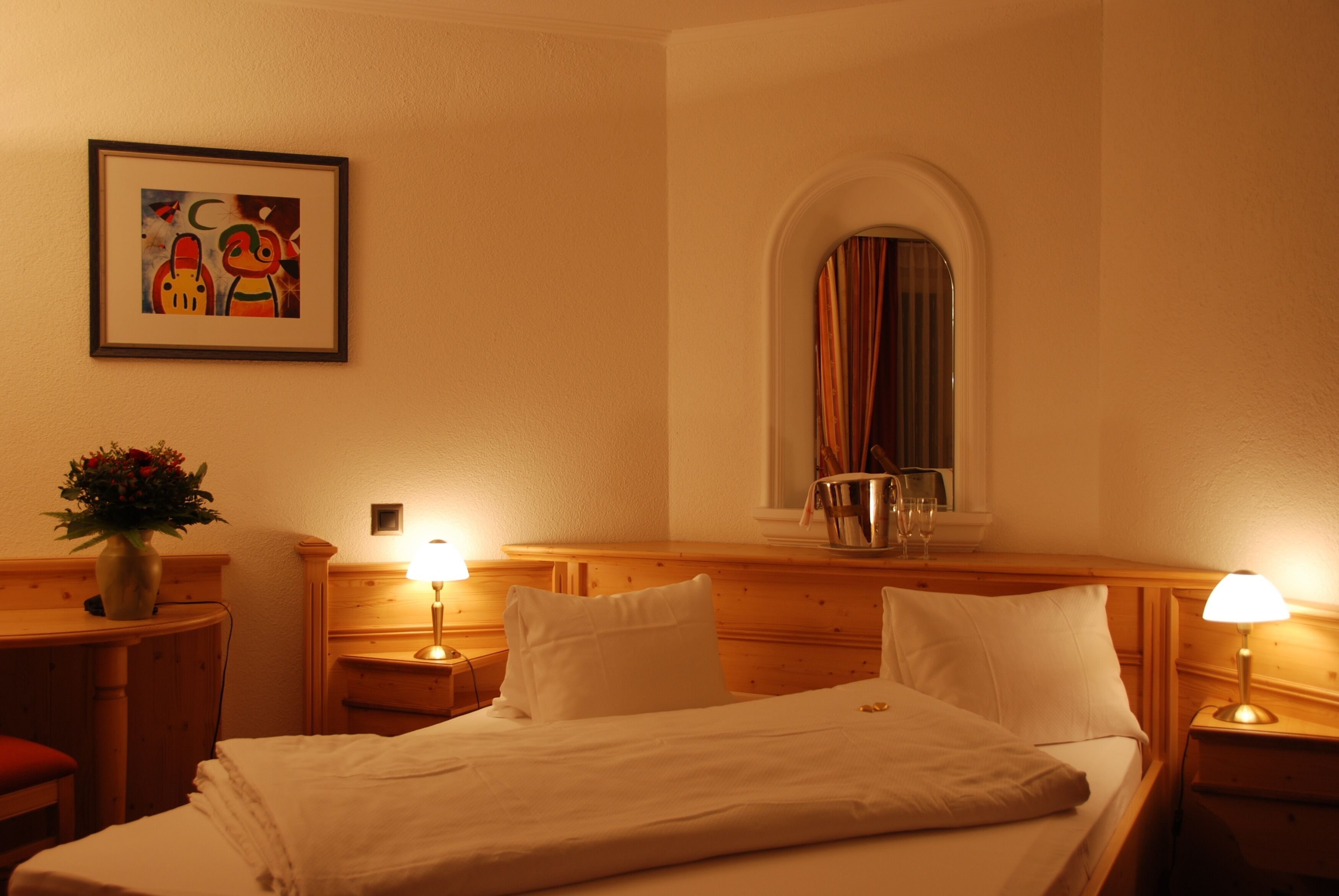 A hotel room with a bed, bedside lamps, a framed picture, a vase with flowers, and a window with curtains.