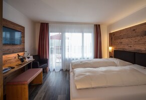 Habitación de hotel con cama, escritorio con silla, televisor montado en la pared y ventana con cortinas.