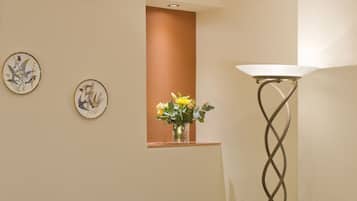A hotel room with a bed, two chairs, a small table with fruit, a vase with flowers, and a tall floor lamp.