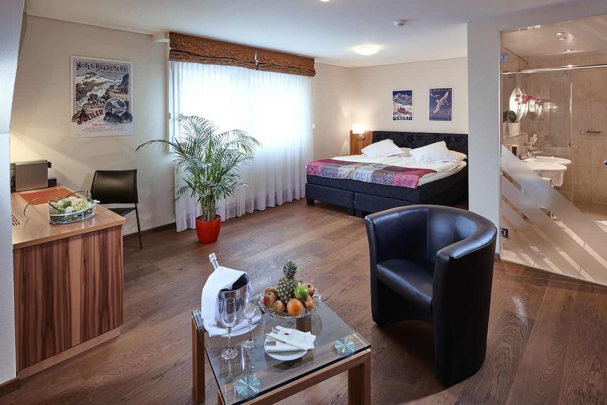 A hotel room with a bed, a desk with a plant, a chair, a glass table with a fruit bowl, and a bathroom with a shower.