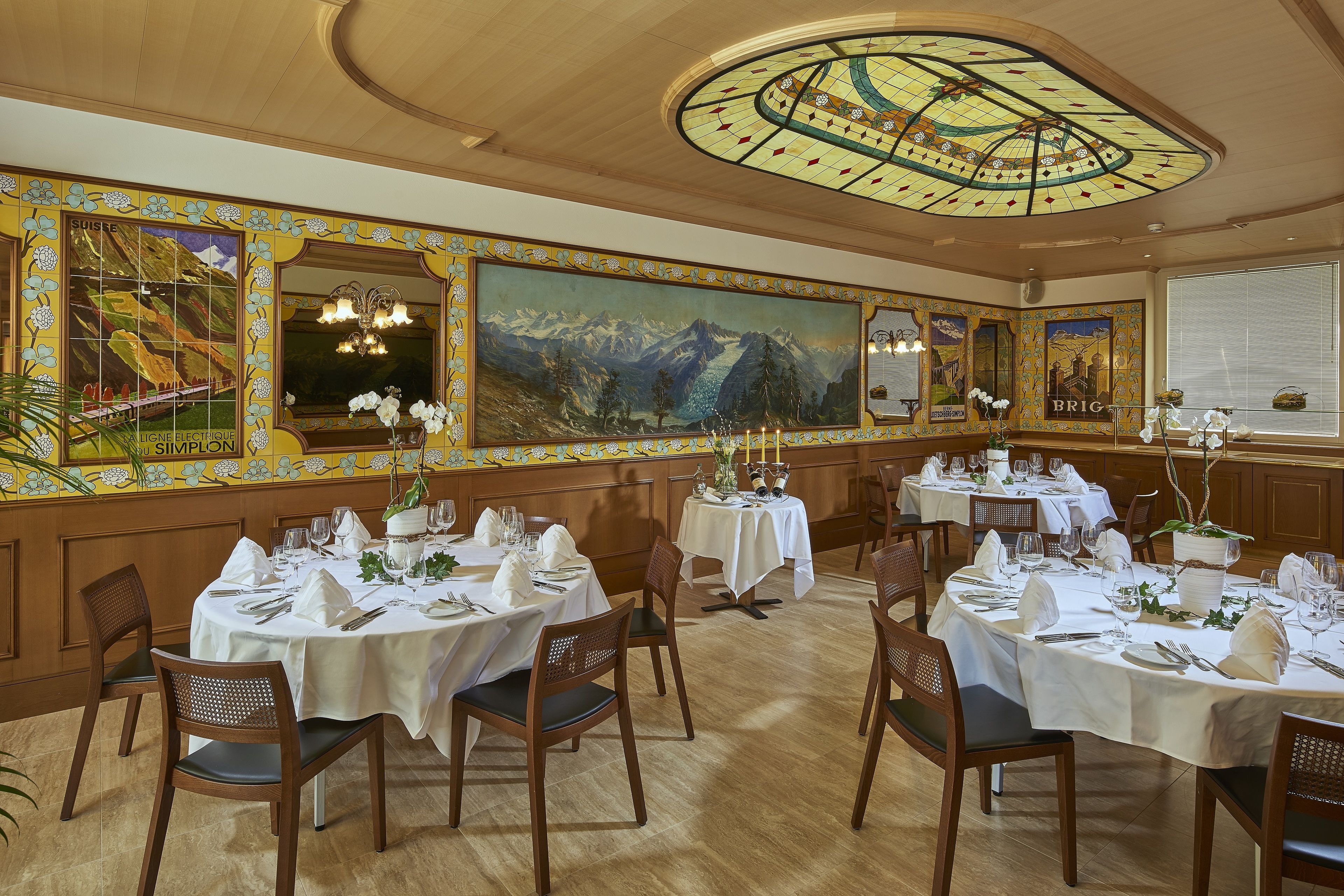 A dining area with round tables set for a meal, wooden chairs, and a large painting of mountains on the wall.