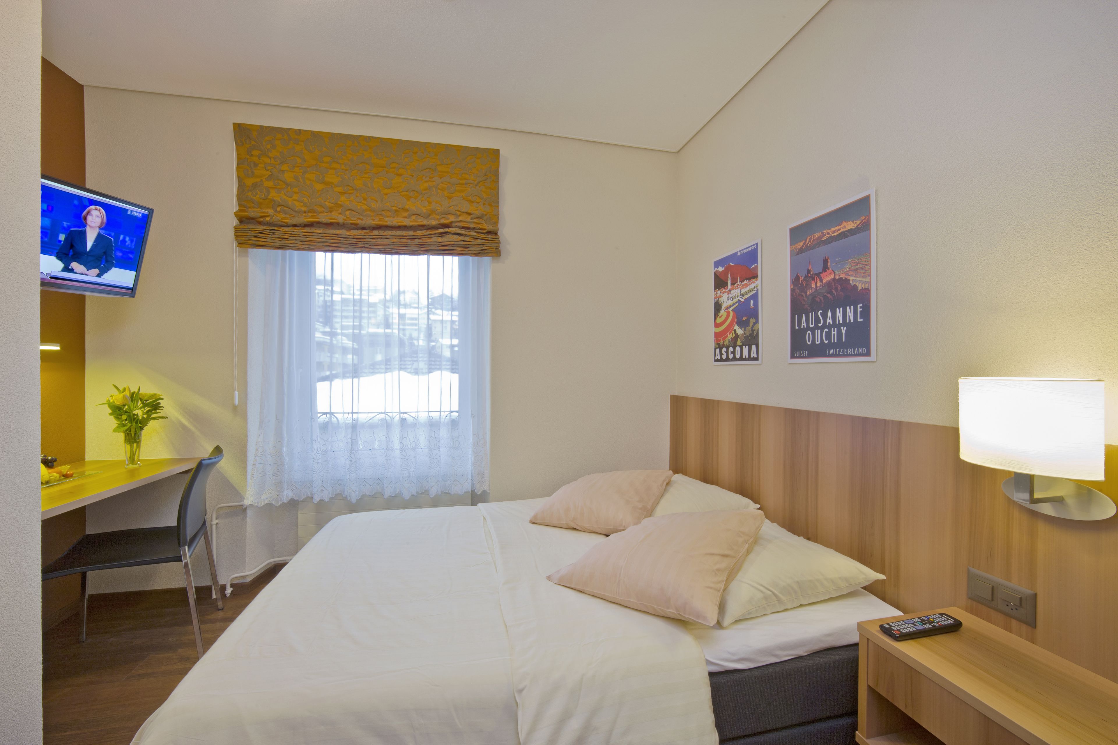 Ein Hotelzimmer mit Bett, Schreibtisch mit Stuhl, Fernseher, Fenster mit Vorhängen und zwei Bildern an der Wand.