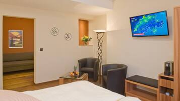 A hotel room with a bed, two chairs, a small table with fruit, a wall-mounted TV displaying a weather map, and a decorative poster.