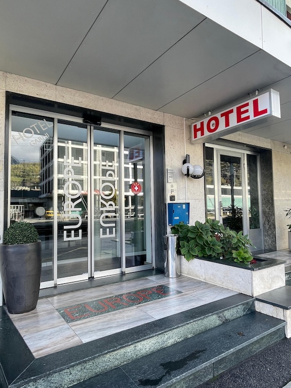 A hotel entrance with a glass door, a sign reading 'HOTEL,' and a small planter with green plants. - Hotel Europe Garni (Brig)