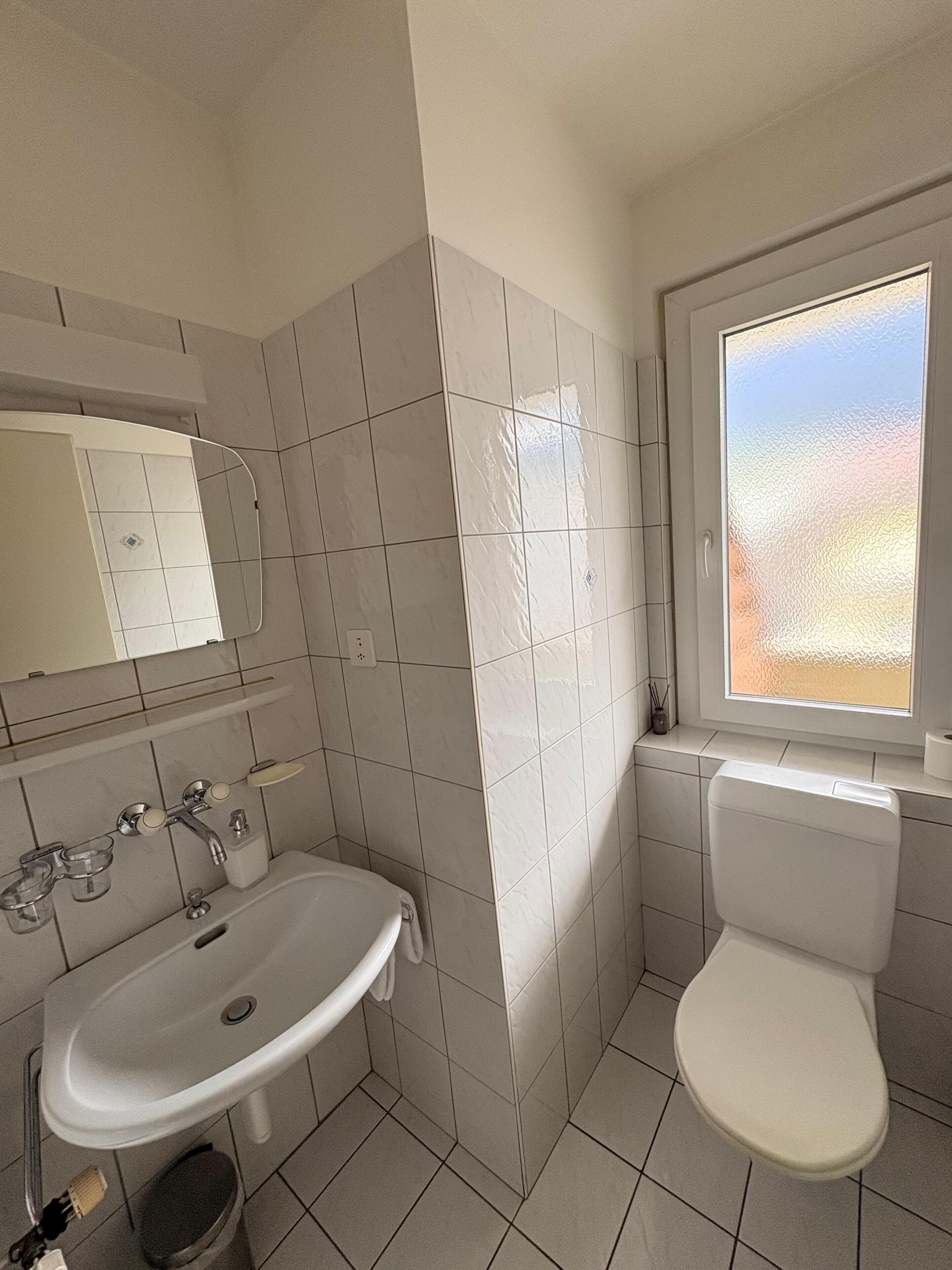 A bathroom with a sink, mirror, toilet, and window.