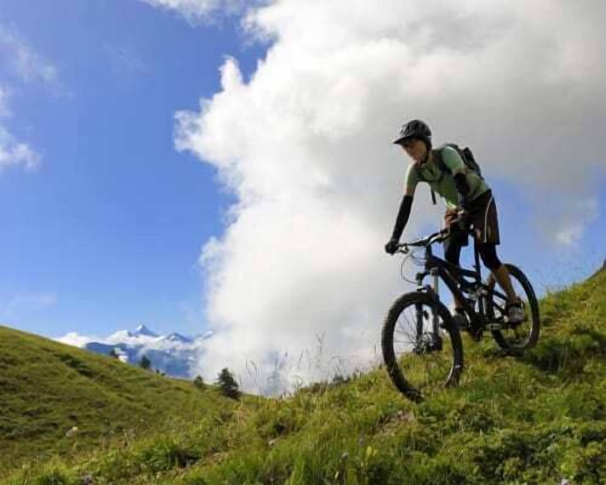 Una persona montando una bicicleta de montaña en una colina cubierta de hierba, con cielo despejado y nubes al fondo.