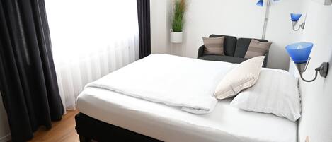 Cama bem arrumada com edredom branco e almofadas, luminária de cabeceira, planta em vaso e sofá com travesseiros em um quarto moderno com piso de madeira.