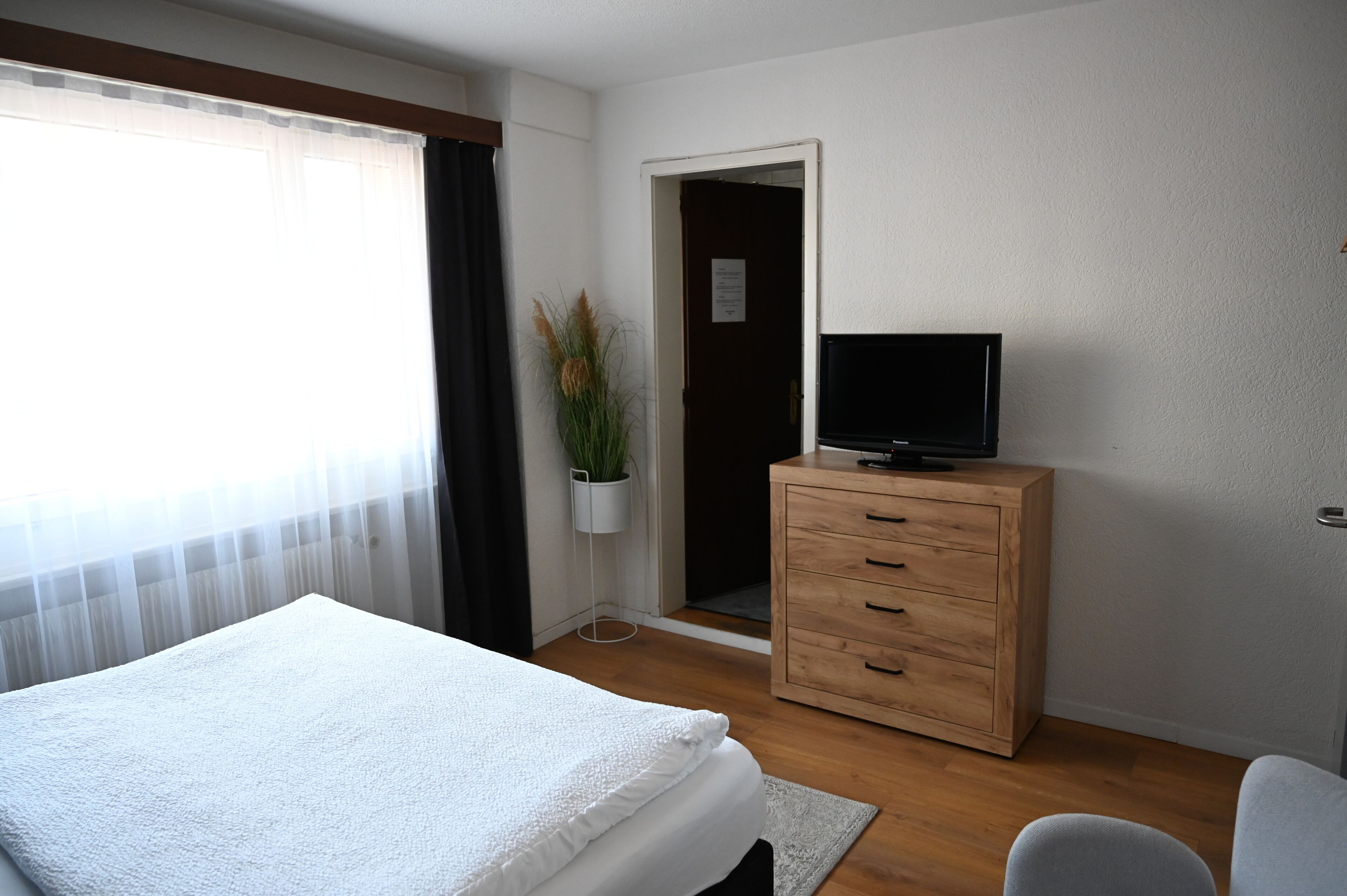 A hotel room with a bed, a wooden dresser, a television, a potted plant, and a door leading to another room.