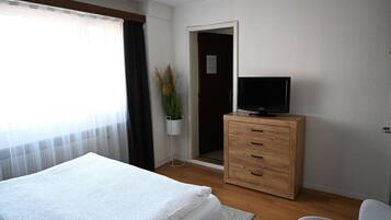 A hotel room with a bed, a wooden dresser, a television, a potted plant, and a door leading to another room.