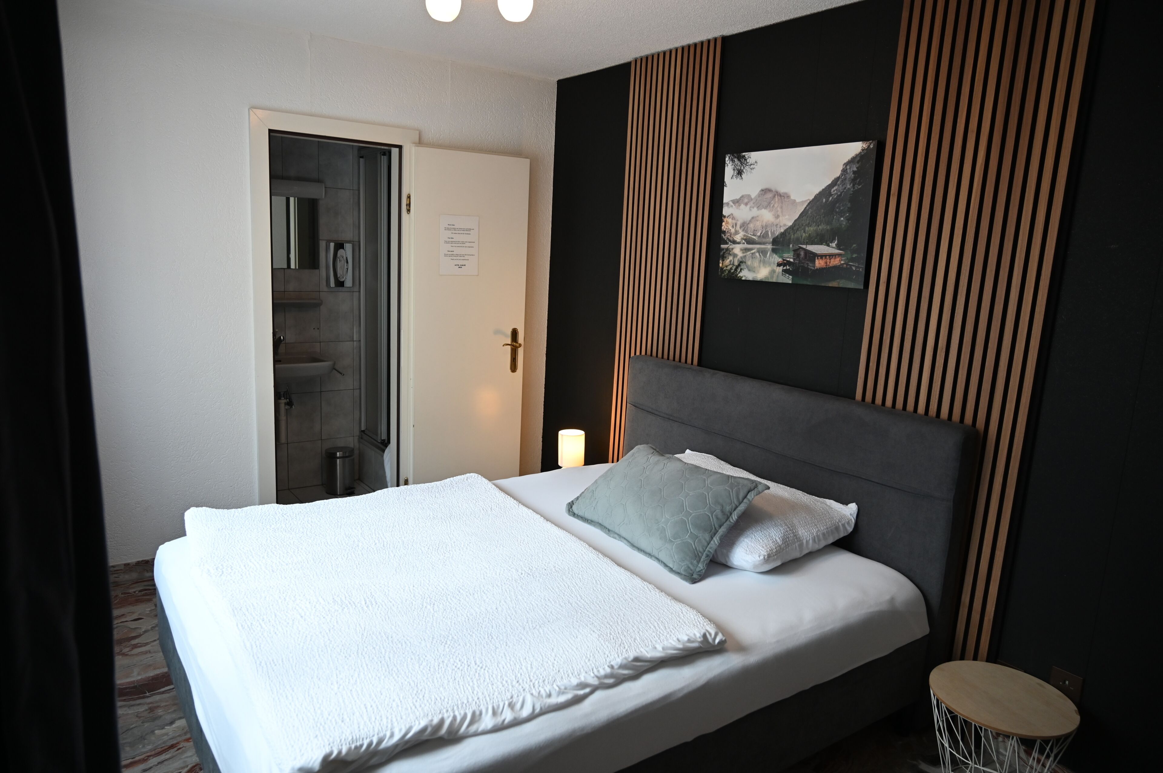 A hotel room with a bed, a bedside table, a nightstand, a wall-mounted picture, and a door leading to a bathroom.