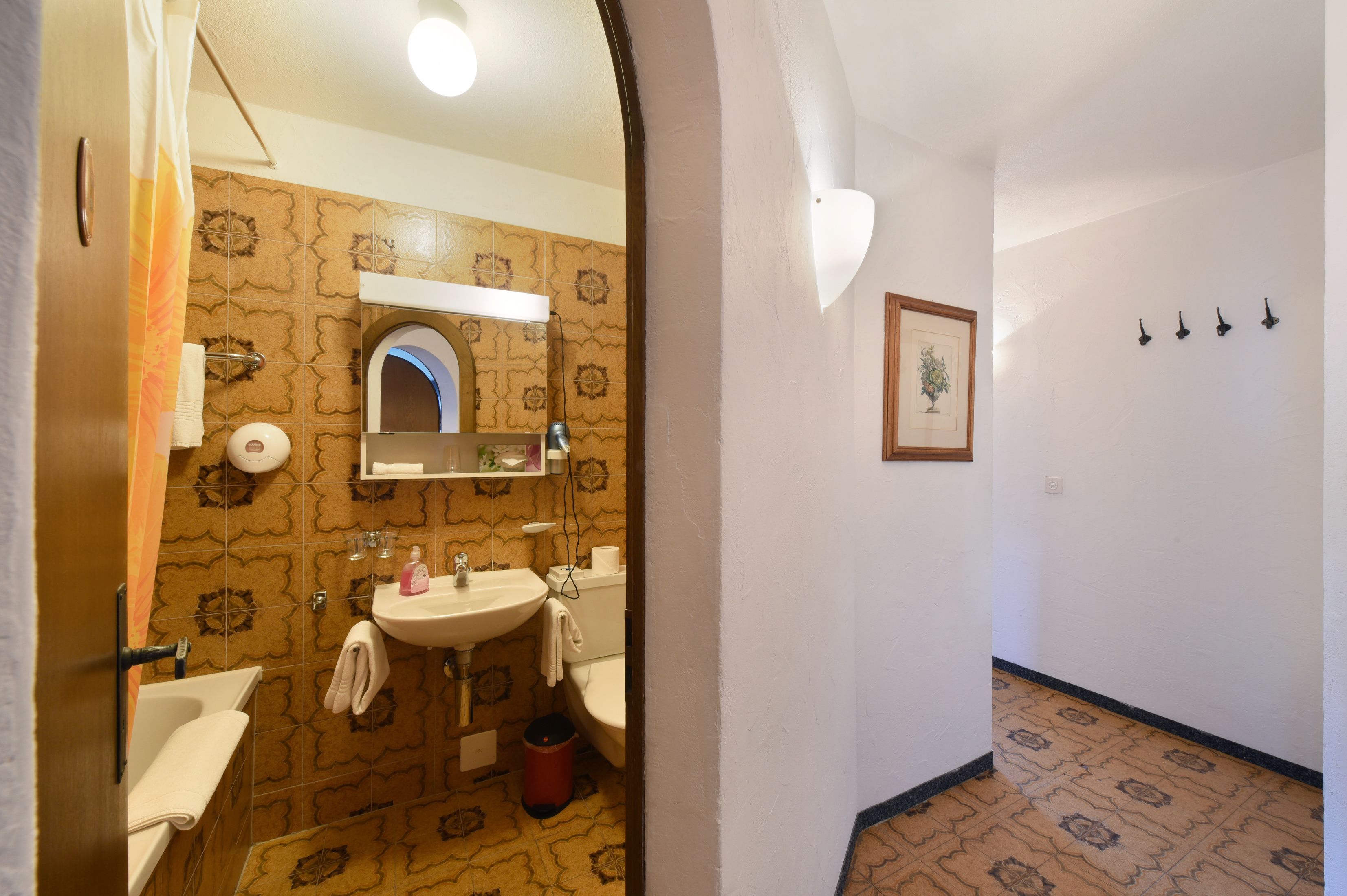 Un baño con lavamanos, espejo e inodoro. Hay una cortina de ducha, un perchero y un cuadro en la pared.