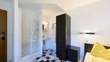 Habitación de hotel con piso a cuadros, ducha, lavamanos, cama con almohadones amarillos y un mueble oscuro.
