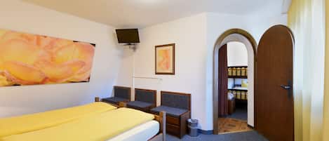 Quarto de hotel com uma cama, uma escrivaninha, uma cadeira, uma televisão presa à parede e uma porta que dá para outro espaço.