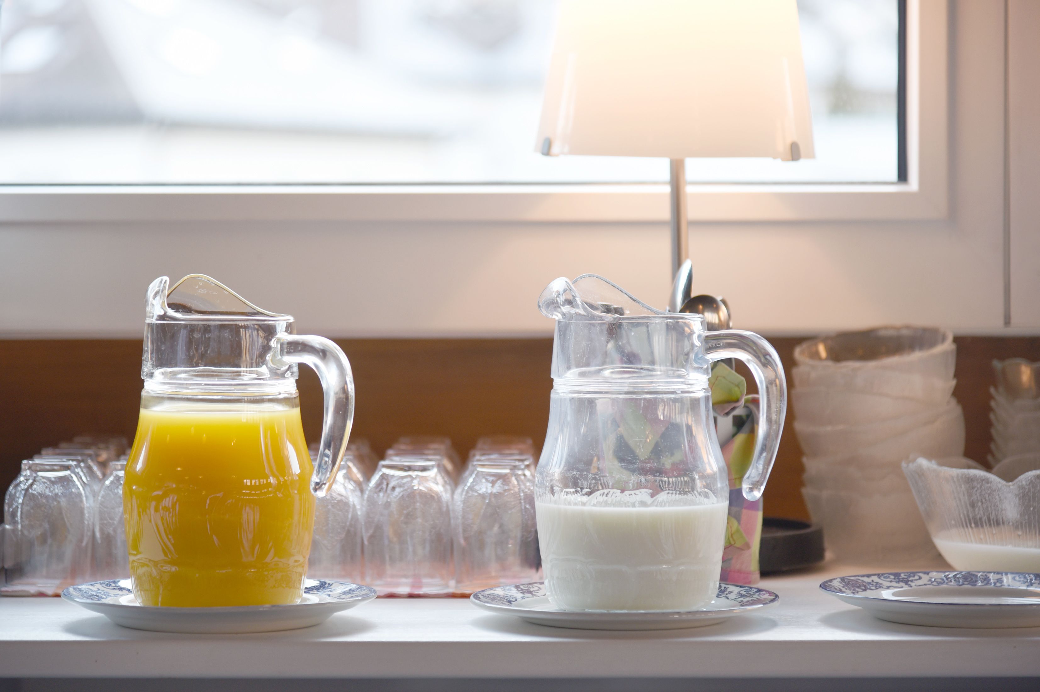 Deux carafes en verre sont posées sur une table : l’une contient du jus d’orange, l’autre du lait. On aperçoit une fenêtre et une lampe en arrière-plan.