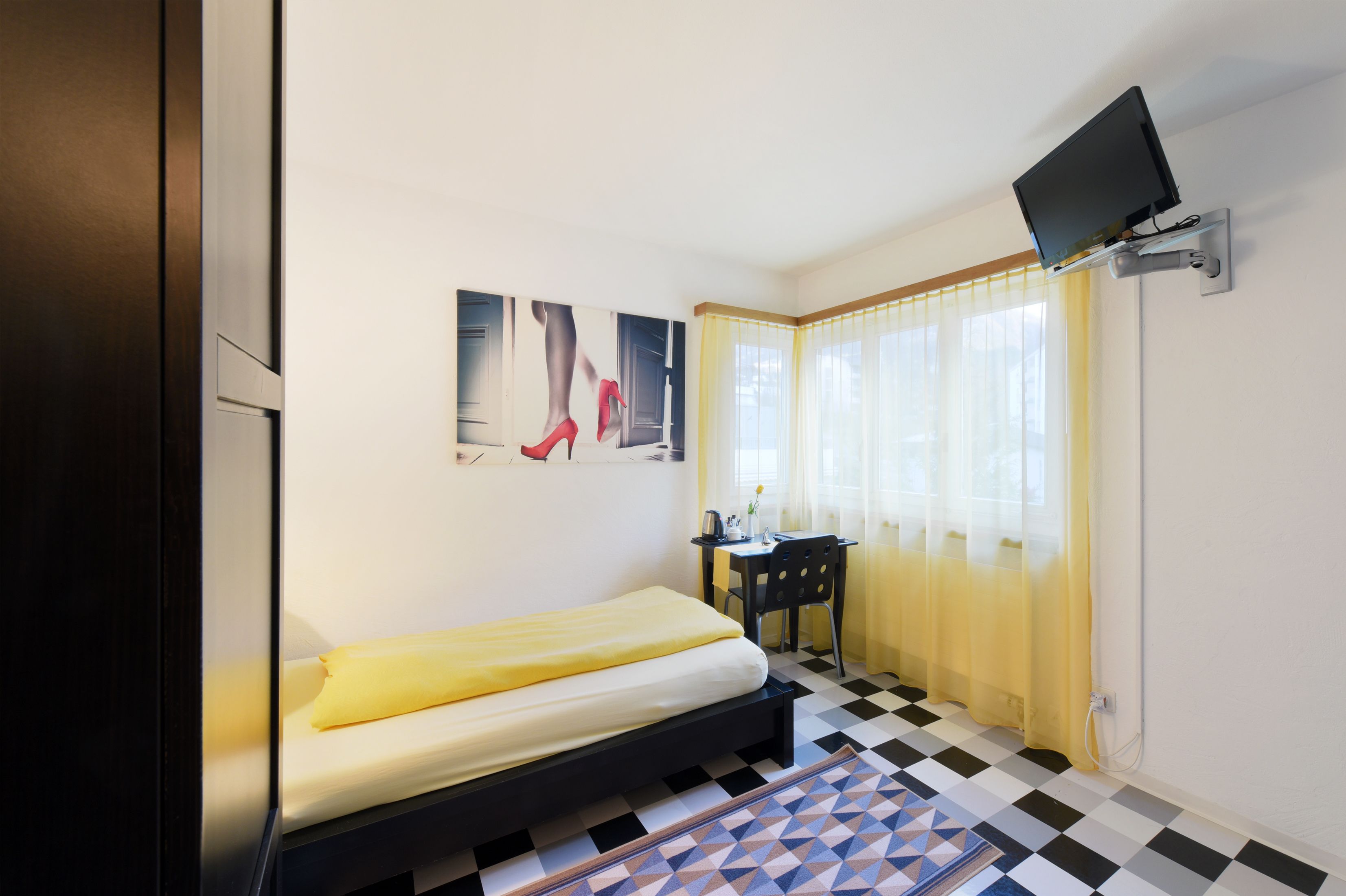 Ein Hotelzimmer mit Bett, Schreibtisch, Stuhl, einem an der Wand befestigten Fernseher und einem Fenster mit Vorhängen.