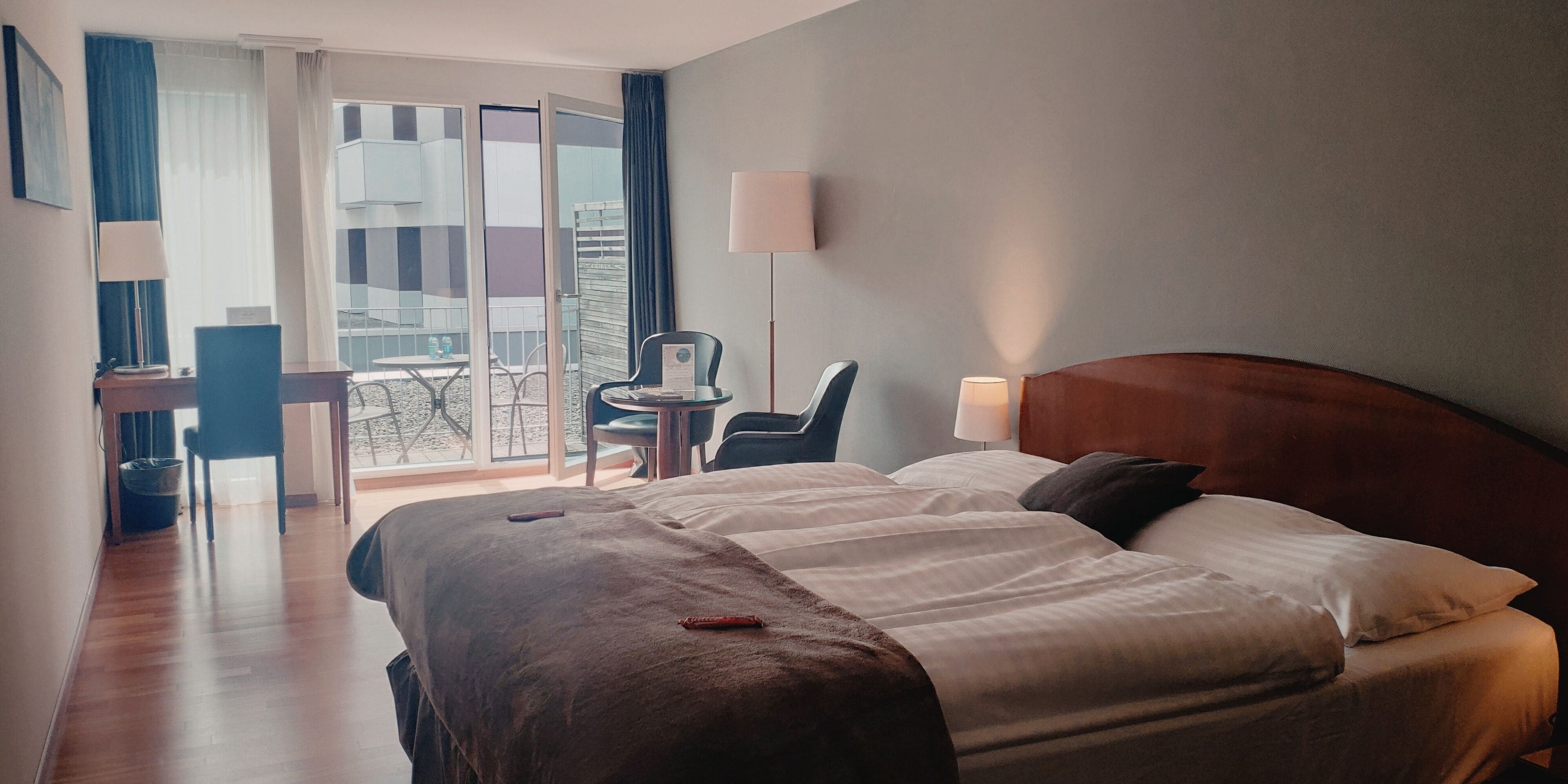 A hotel room with a large bed, a desk, and a chair. There is a balcony with a view of buildings.