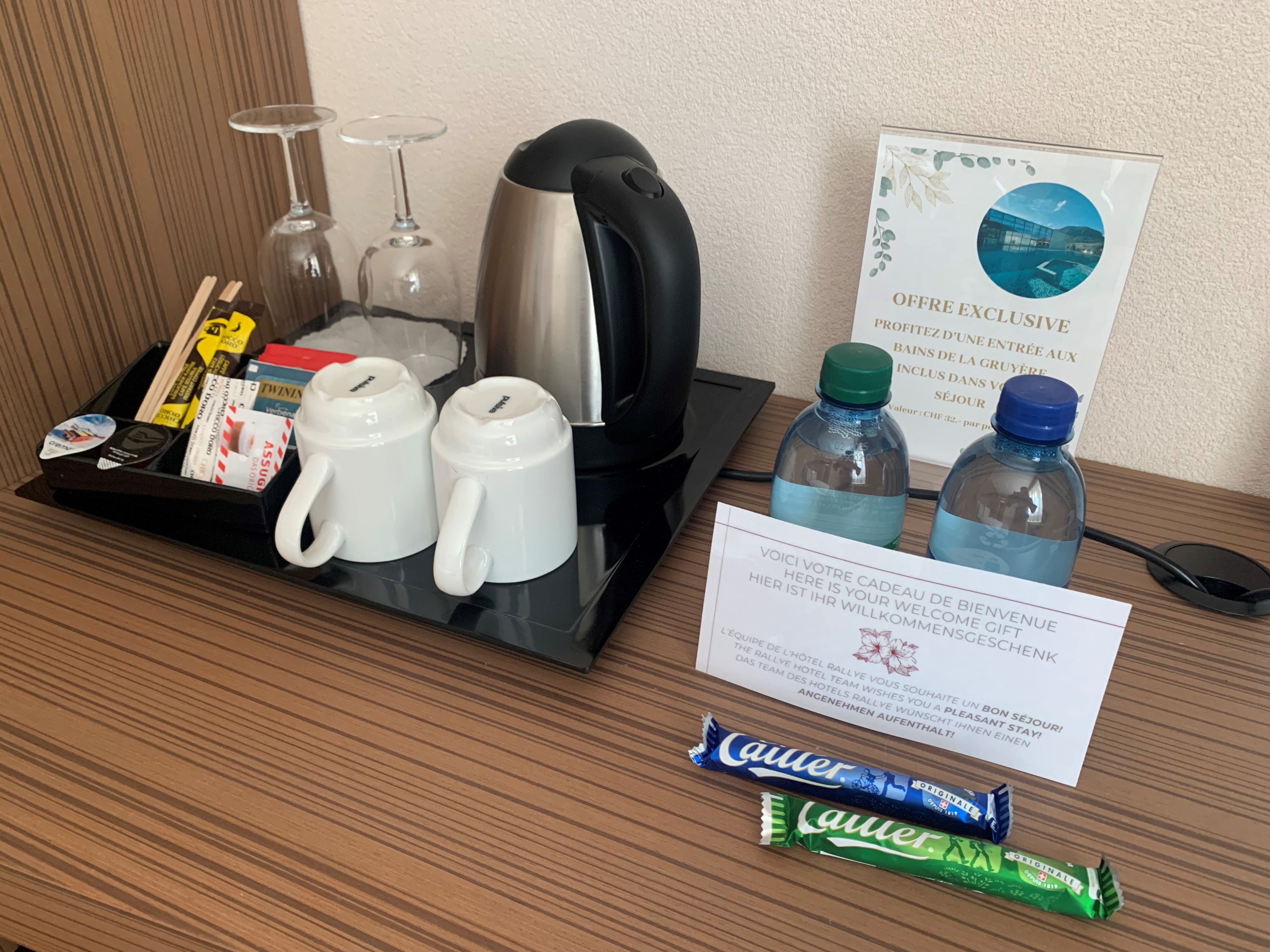 Zona de bienvenida en una habitación de hotel, con una bandeja de servicios que incluye un hervidor, vasos, una botella de agua y una tarjeta de bienvenida.
