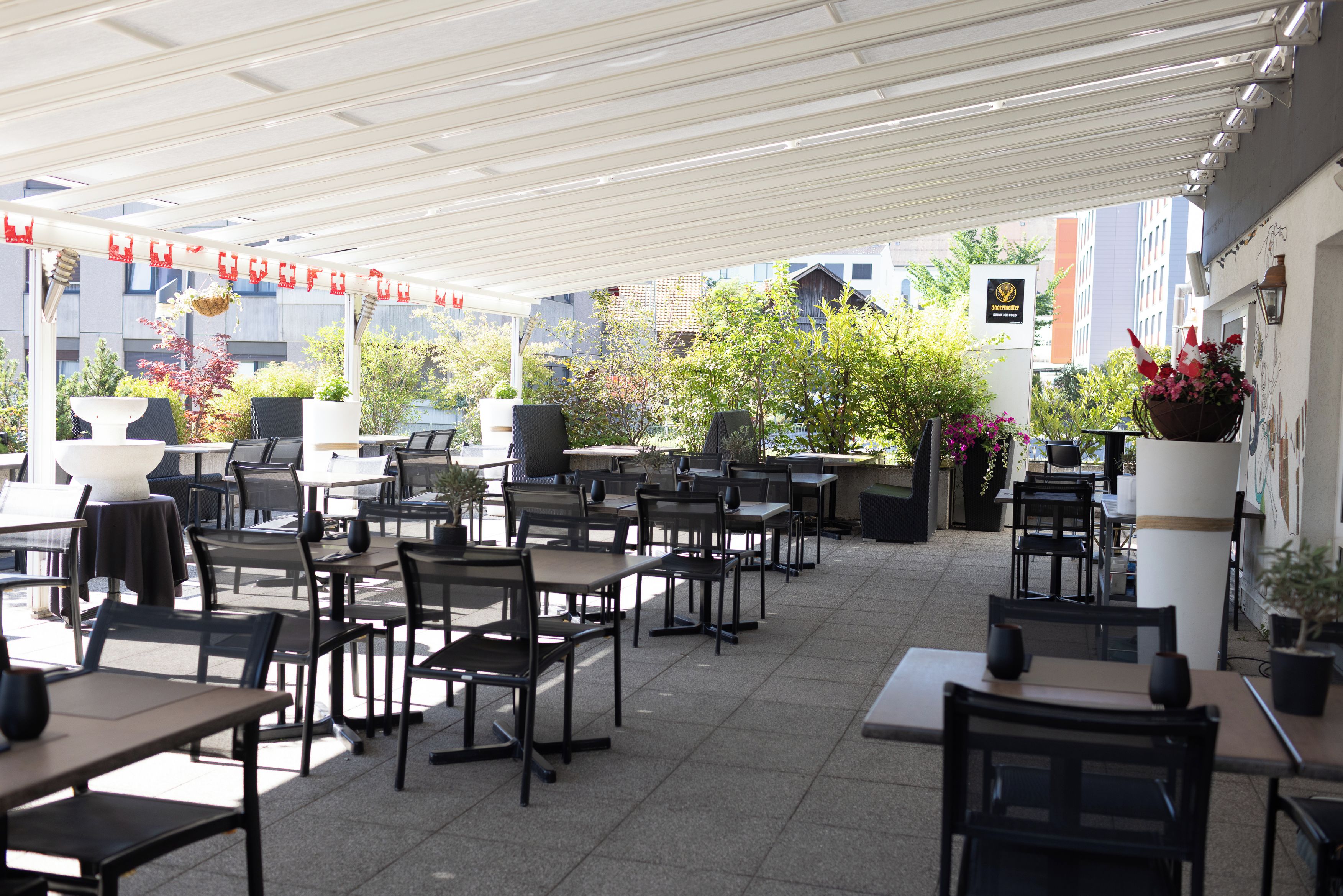 An outdoor café area with tables and chairs, potted plants, and a view of buildings in the background.
