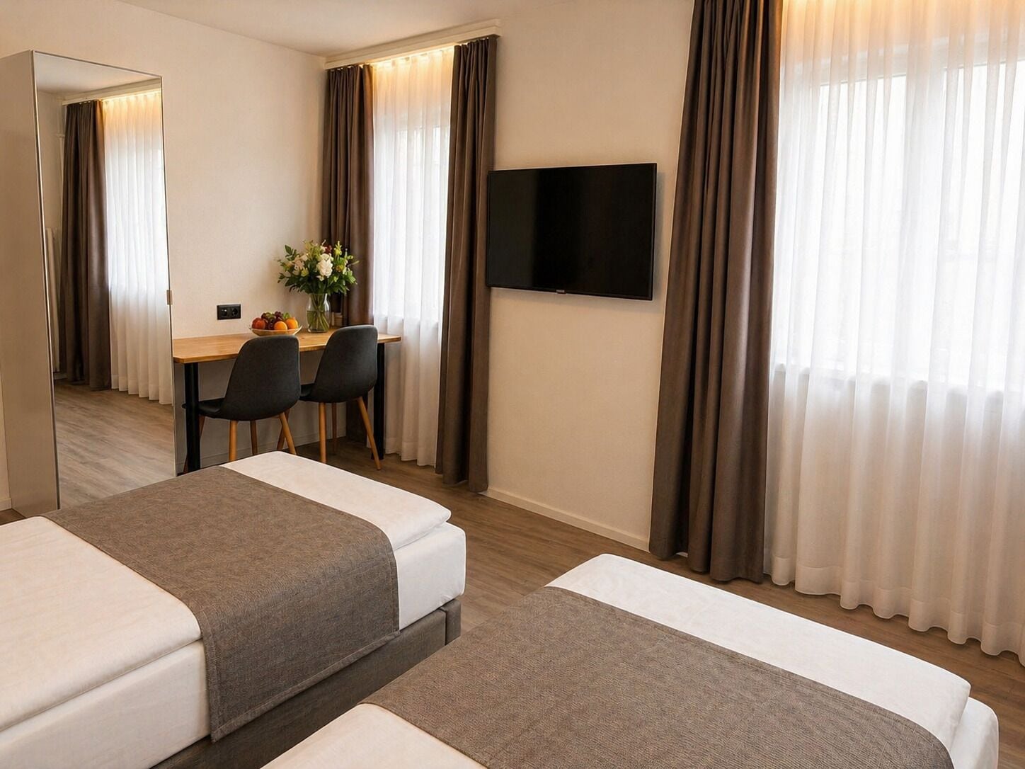 A hotel room with two beds, a desk with chairs, a flat-screen TV, and a large mirror.