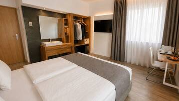 A hotel room with a bed, a desk with a chair, a TV, a mirror, a wardrobe, and a window with curtains.