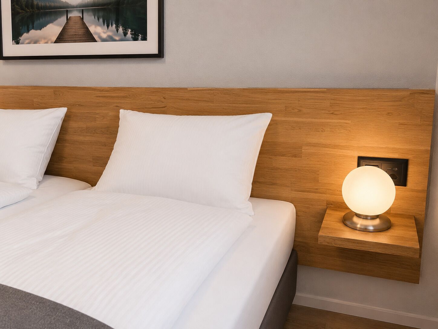 A neatly made bed with white linens, a wooden headboard, a bedside table with a round lamp, and a framed picture above the bed.