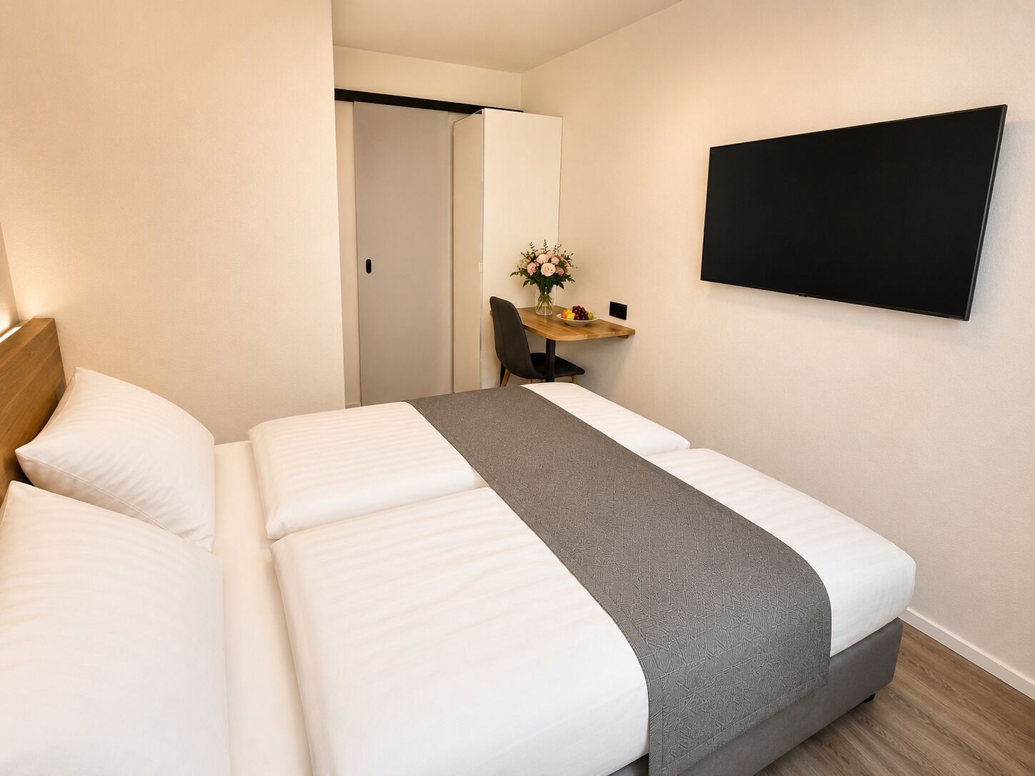 Una habitación de hotel con una cama, un escritorio con silla, un florero con flores y un televisor montado en la pared.