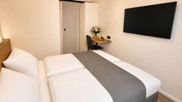 Una habitación de hotel con una cama, un escritorio con silla, un florero con flores y un televisor montado en la pared.