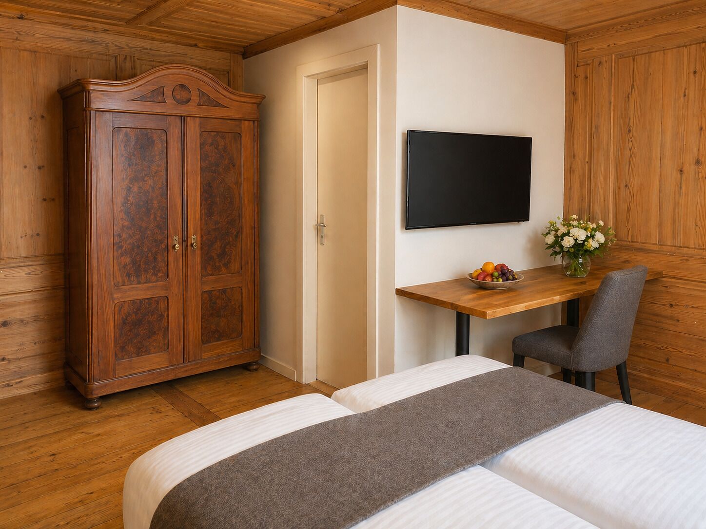 A bedroom with a wooden wardrobe, a desk with a chair, a bed, and a TV on the wall.
