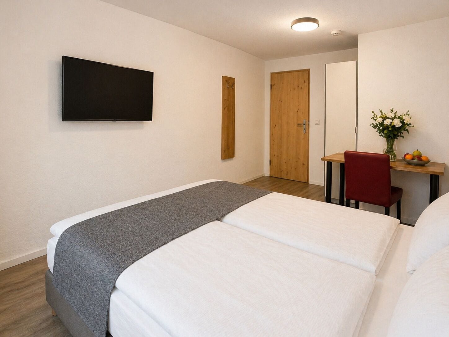 Habitación de hotel con cama, escritorio con silla, televisor montado en la pared y puerta que da a otra habitación.