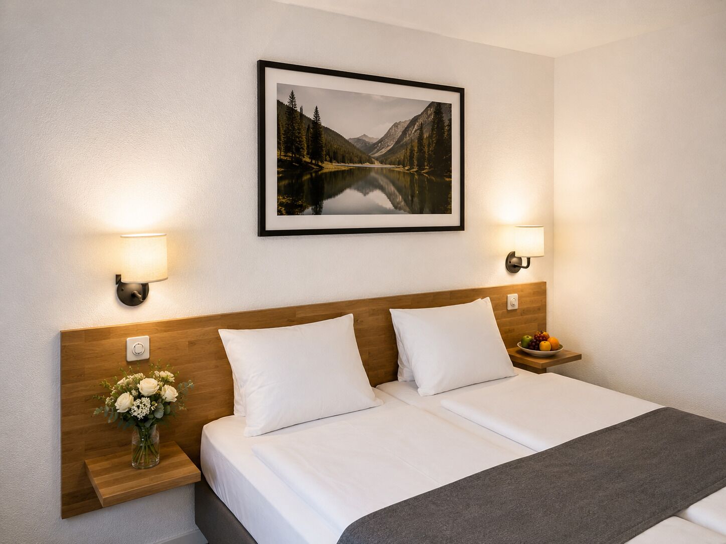 Ein Hotelzimmer mit einem hölzernen Kopfteil, zwei weißen Kissen, einem gerahmten Landschaftsbild, Nachttischlampen, einem Nachttisch mit Blumenvase und einer Obstschale.
