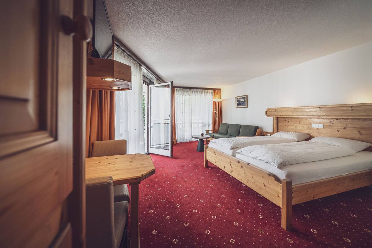 A hotel room with two single beds, a wooden headboard, a small table, a chair, and a view of the outdoors through a sliding door.