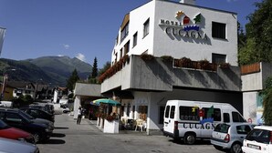 Hotelli nimeltä »Hotel Cucagna», jossa on moderni muotoilu, pysäköintialue ja vuoristomaisema taustalla.