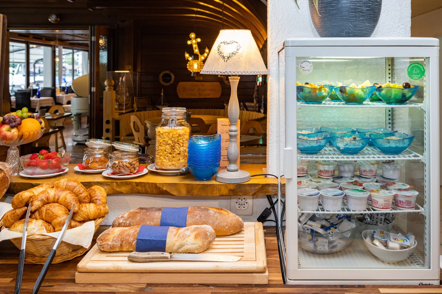 A breakfast buffet with various food items, including bread, pastries, and a refrigerator stocked with yogurt and fruit bowls.
