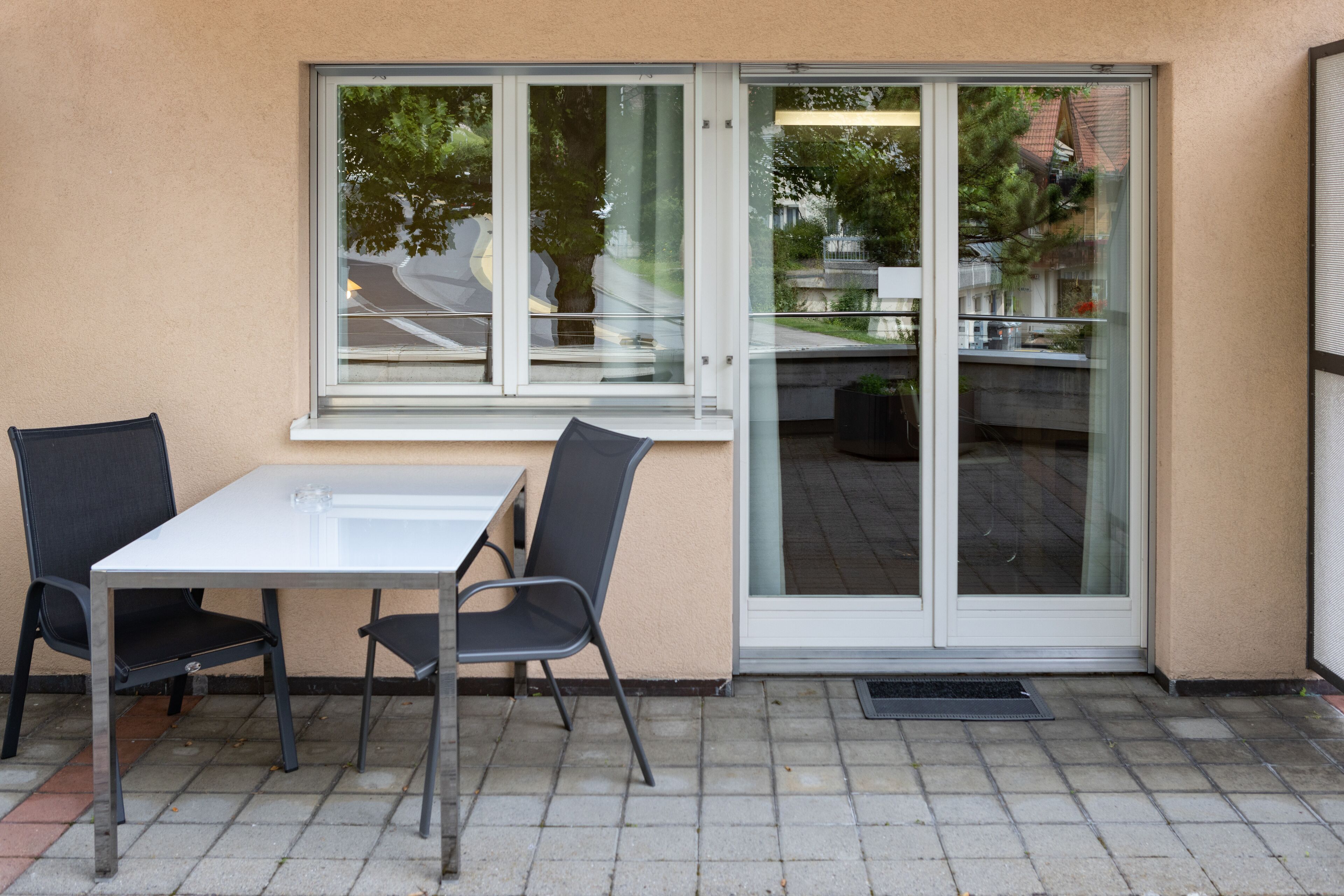 Eine Terrasse im Freien mit einem weißen Tisch und zwei Stühlen, einer Glasschiebetür und Blick auf die Straße und Grünflächen.