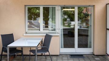 Eine Terrasse im Freien mit einem weißen Tisch und zwei Stühlen, einer Glasschiebetür und Blick auf die Straße und Grünflächen.