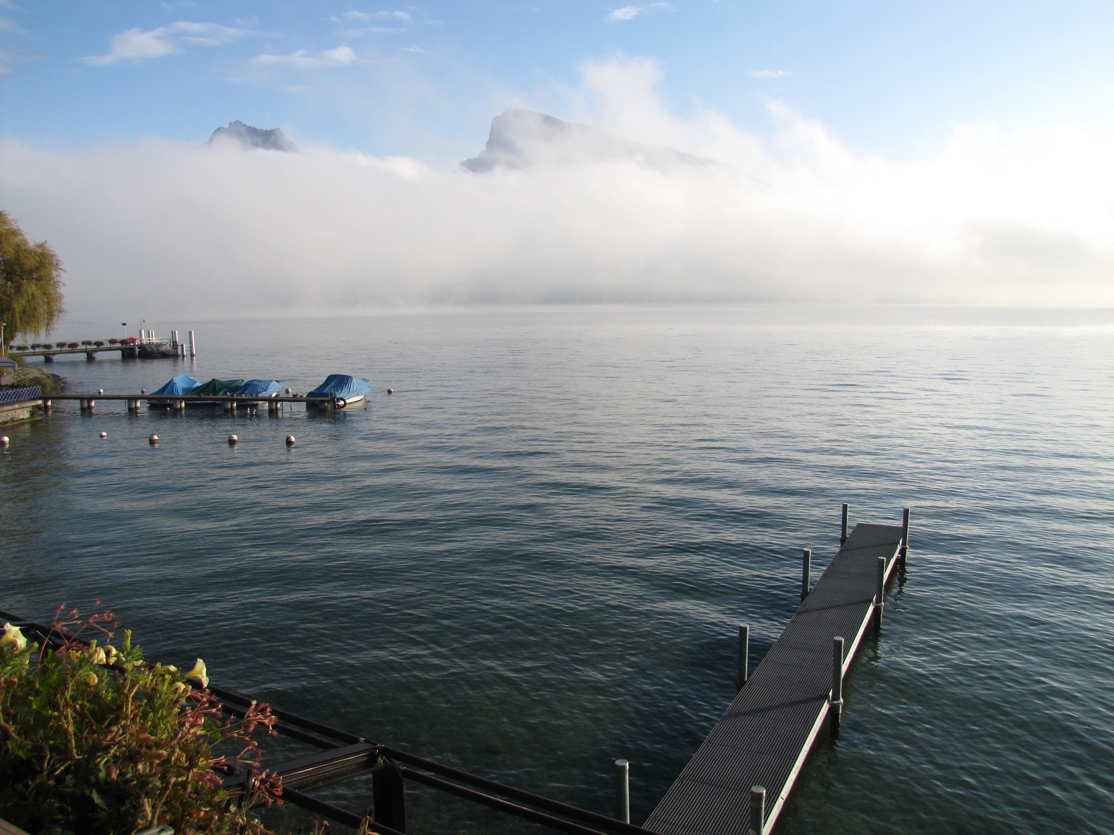 A calm lake with a dock extending into the water, covered boats, and a distant mountain range partially obscured by clouds.