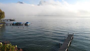 A calm lake with a dock extending into the water, covered boats, and a distant mountain range partially obscured by clouds.