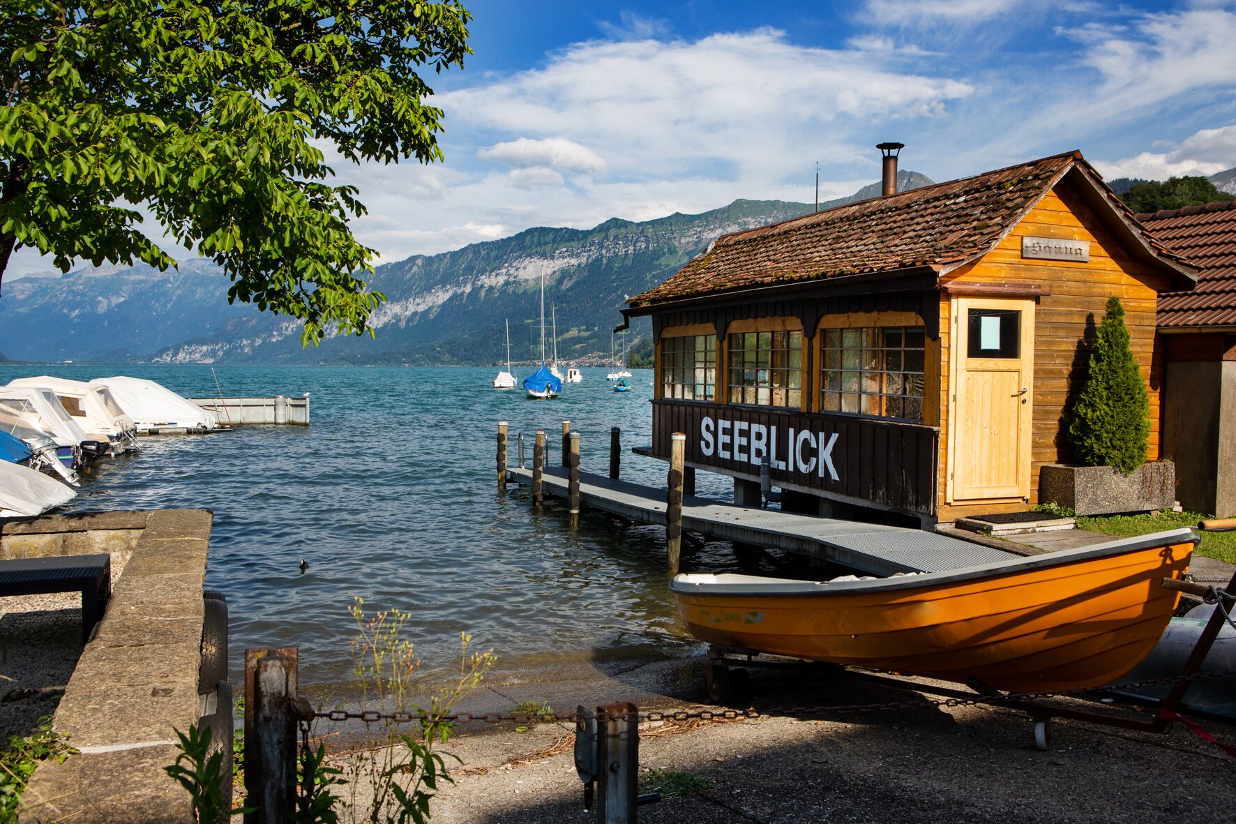 A wooden cabin with a sign reading "SEEBLICK" by a calm lake with boats docked.