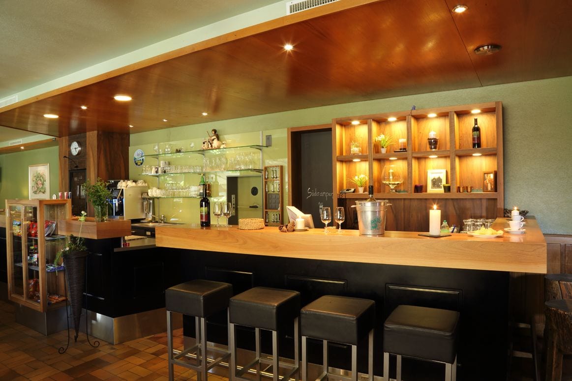 A bar area with a wooden counter, bar stools, shelves with bottles and glasses, and a clock on the wall.