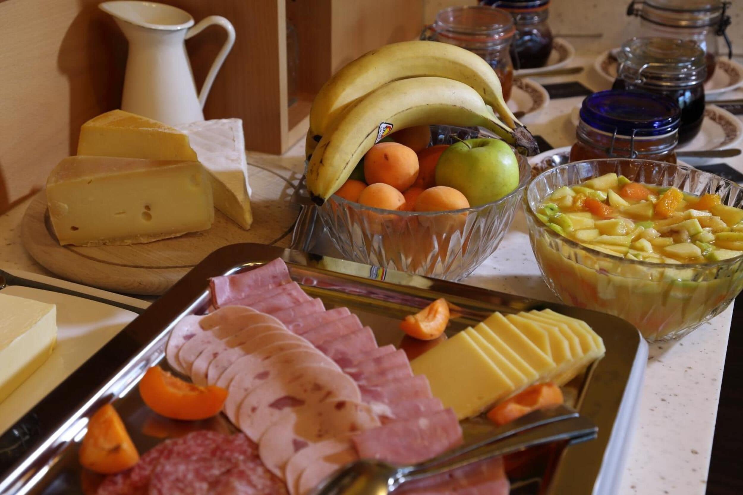 A buffet with cheese, ham, sliced meats, bananas, oranges, apples, and a fruit salad.