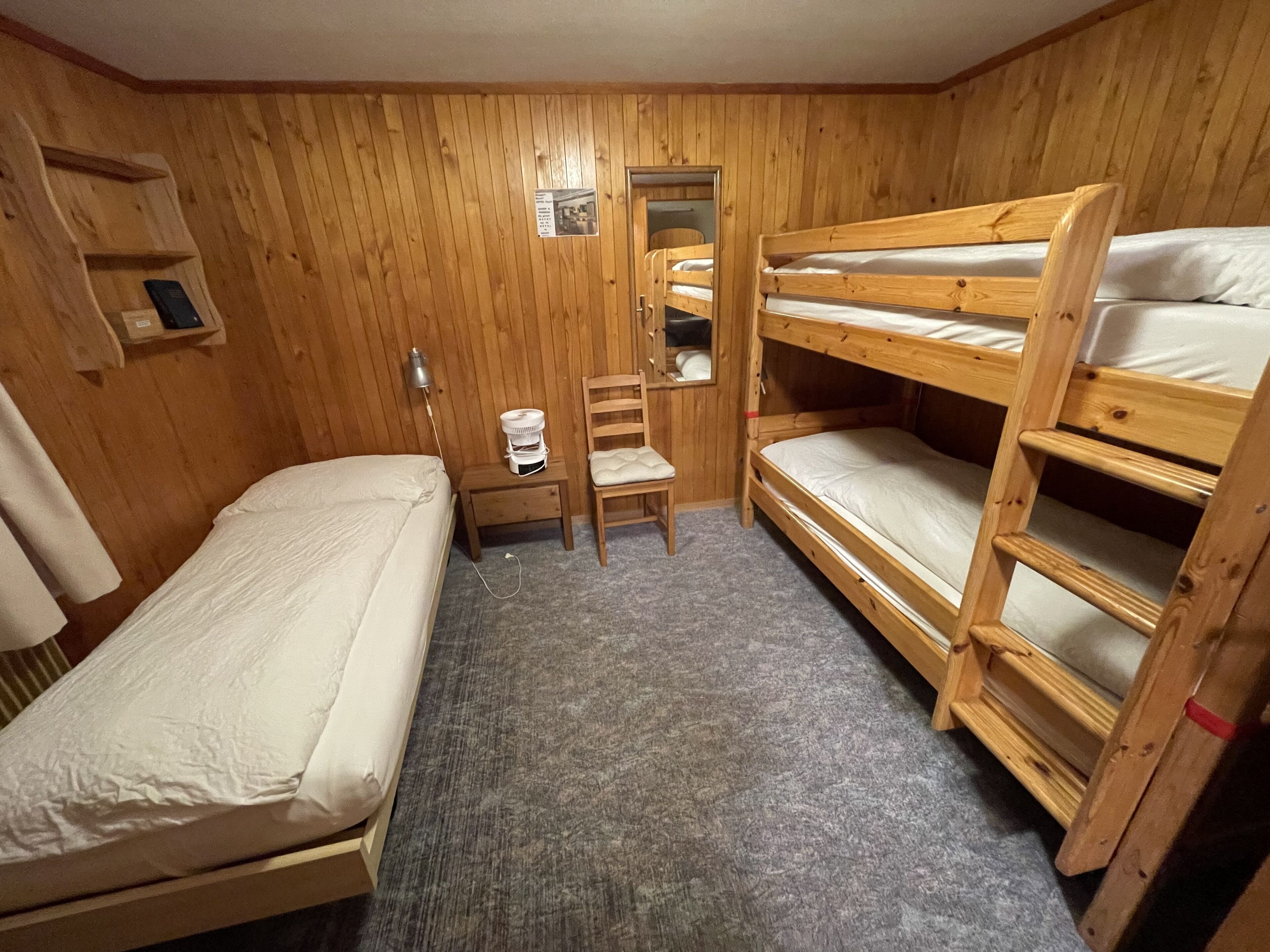 A small, wooden room with bunk beds, a single bed, a chair, a small table, and a mirror.