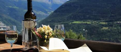 Un libro, dos copas de vino, una botella y un florero con flores sobre una mesa de madera con vista a la montaña.
