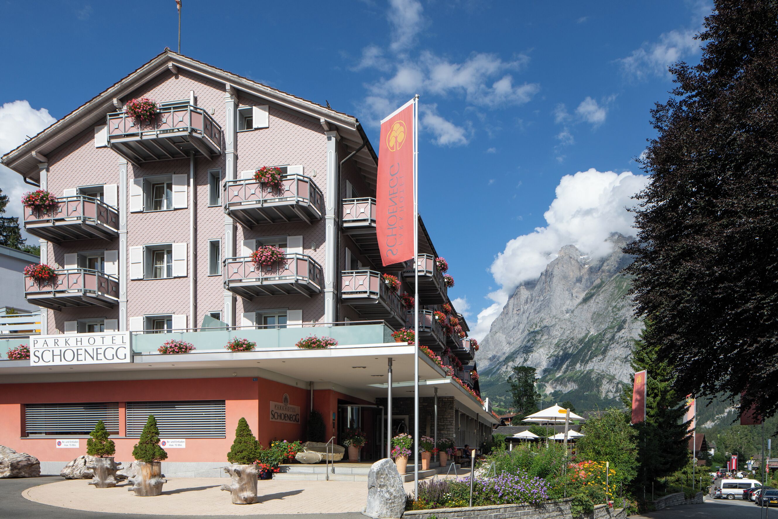 A multi-story hotel with balconies adorned with flowers, a prominent flag, and a mountain backdrop.