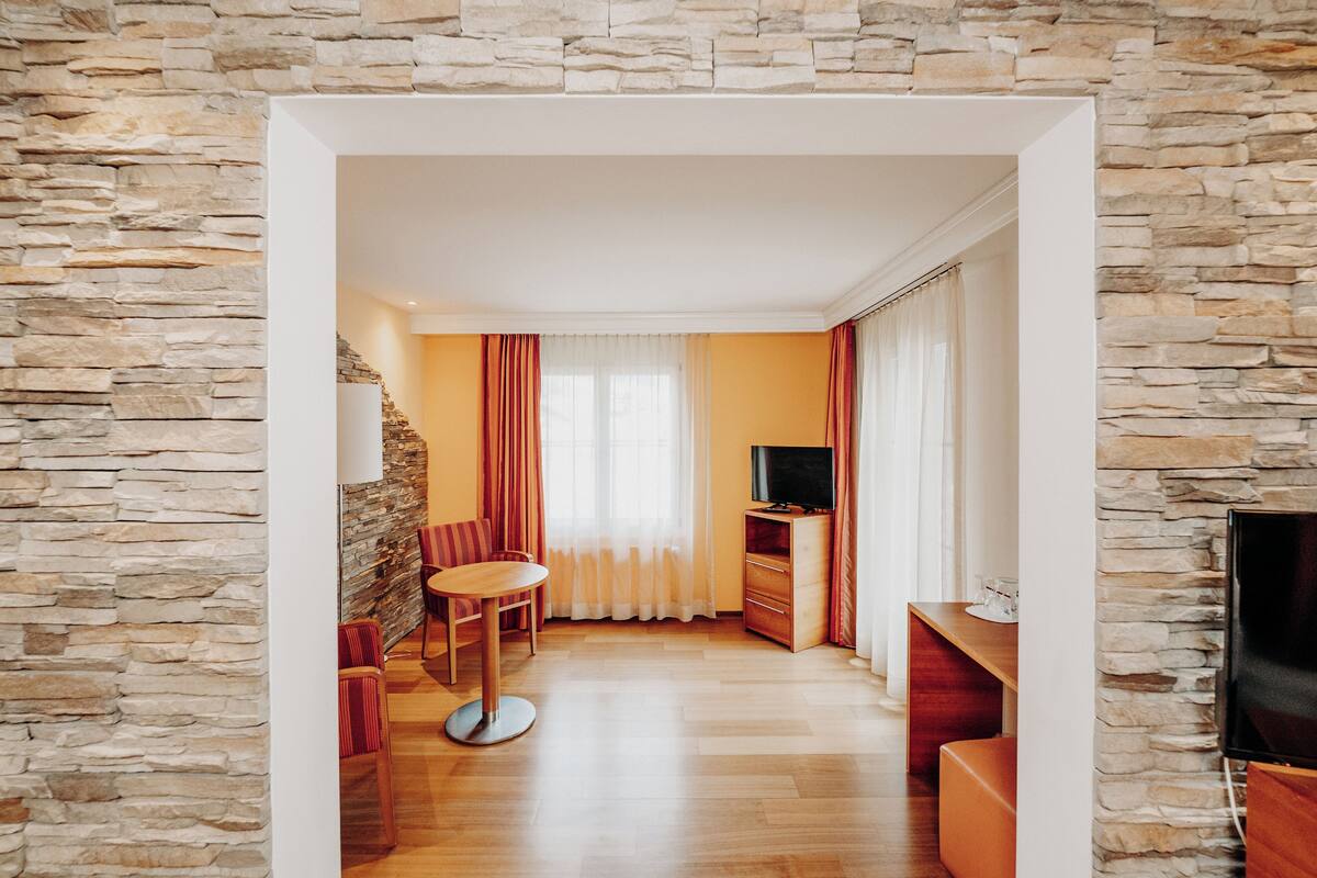 A room with a stone wall, a wooden dining table with chairs, a television, and a window with curtains.