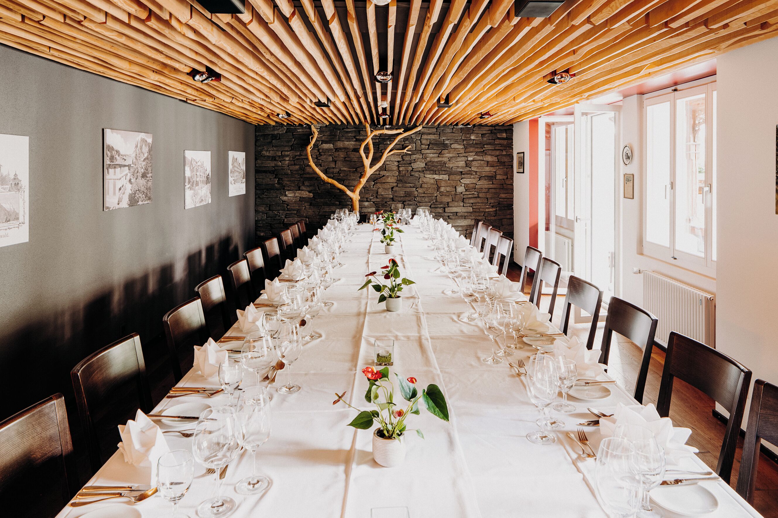 A long dining table set for a formal event, with chairs on both sides, a wooden ceiling, and a stone wall.