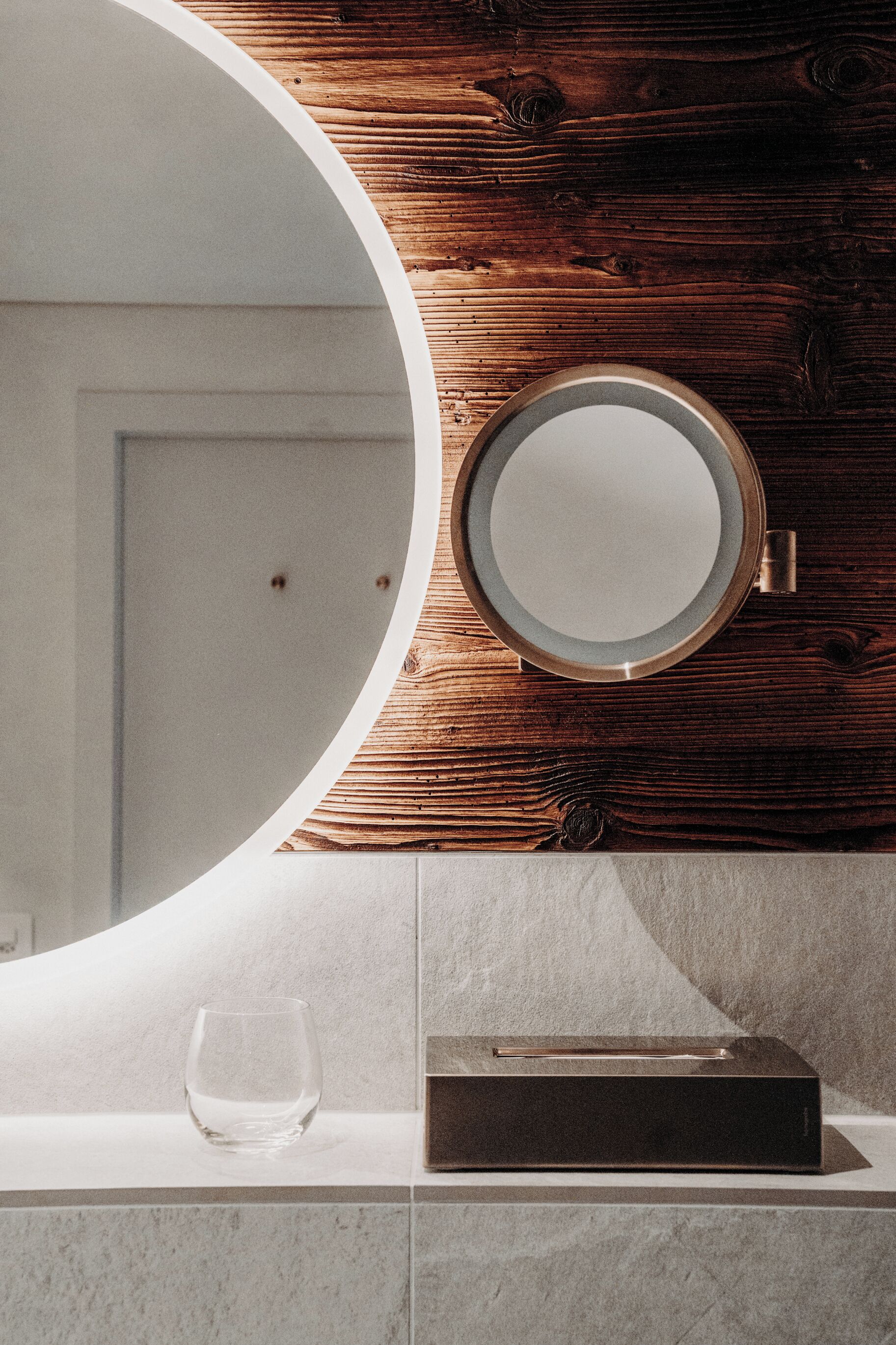 A bathroom with a wooden wall, a round mirror, a glass, and a black box on the counter.