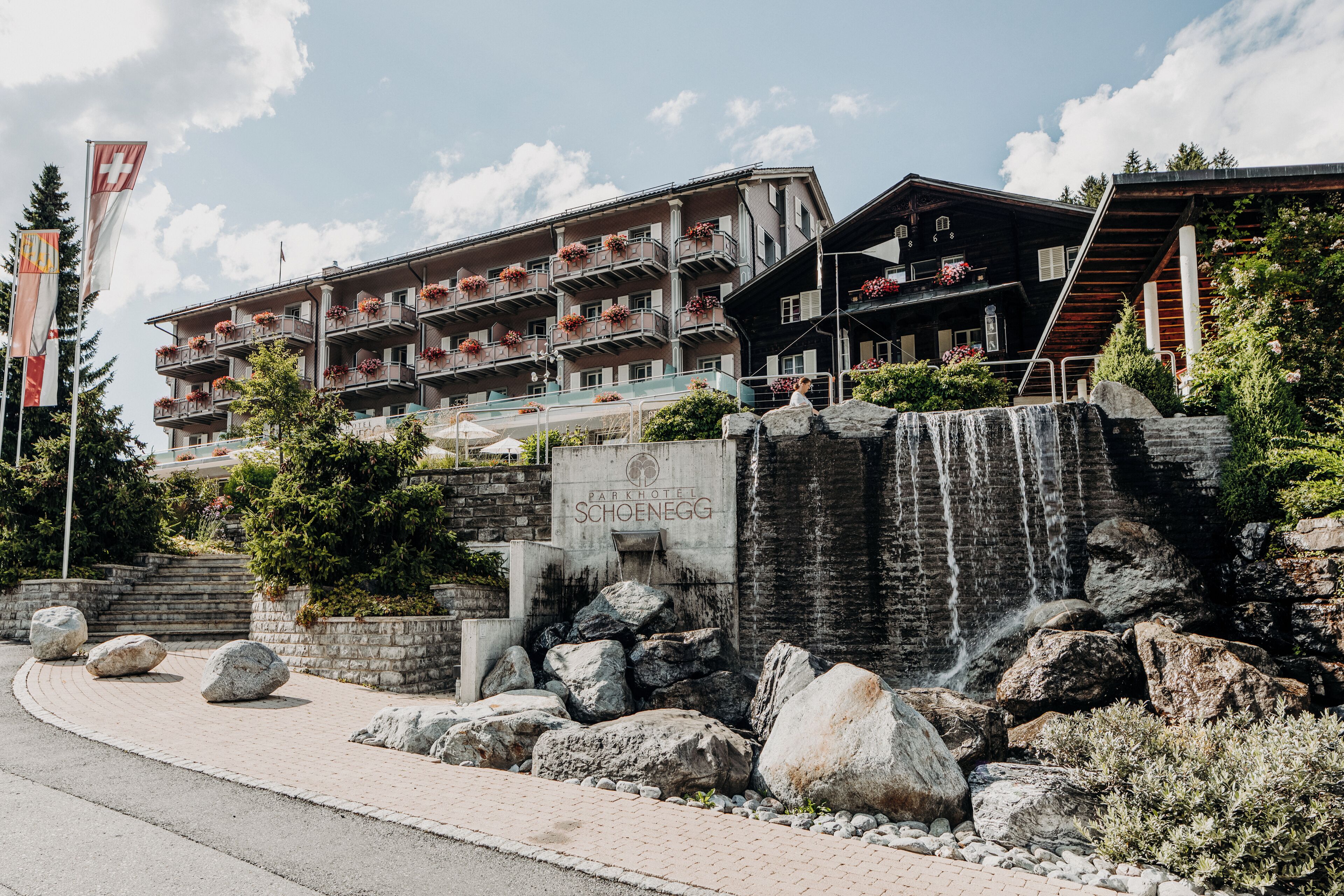 A hotel with multiple balconies adorned with flowers, a sign reading "Parkhotel Schoenegg," and a waterfall feature.